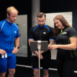 Drei Personen in Sportkleidung stehen neben einer digitalen Waage oder einem Fitnessgerät. Eine Frau rechts zeigt auf den Bildschirm eines Tablets und erklärt den beiden Männern in Sportkleidung, die aufmerksam auf das Display blicken, etwas. Drei Personen in Sportkleidung stehen neben einer digitalen Waage oder einem Fitnessgerät. Eine Frau rechts zeigt auf den Bildschirm eines Tablets und erklärt den beiden Männern in Sportkleidung, die aufmerksam auf das Display blicken, etwas.
