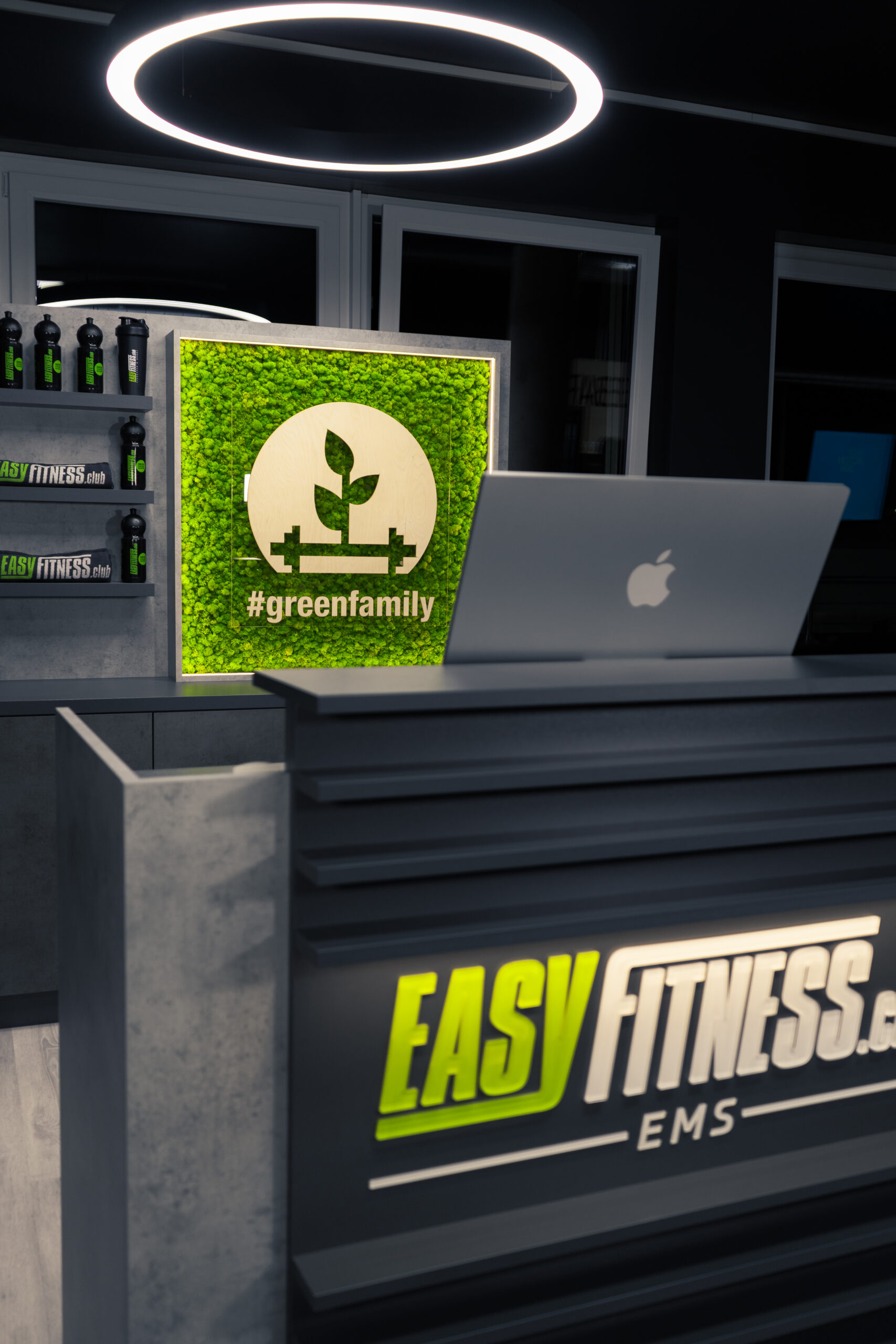 Ein moderner Empfangstresen im Fitnessstudio mit einem Laptop, einem beleuchteten EASY FITNESS EMS-Schild, Wasserflaschen auf einem Regal und einem grünen Wanddisplay mit einer Pflanze und dem Hashtag #greenfamily.