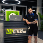 Ein Mann im schwarzen Sportanzug steht lächelnd an der Rezeption eines modernen Fitnessclubs namens EASYFITNESS.club EMS. Im stilvollen, gut beleuchteten Innenraum sind Pflanzen und Fitnessprodukte zu sehen. Ein Mann im schwarzen Sportanzug steht lächelnd an der Rezeption eines modernen Fitnessclubs namens EASYFITNESS.club EMS. Im stilvollen, gut beleuchteten Innenraum sind Pflanzen und Fitnessprodukte zu sehen.