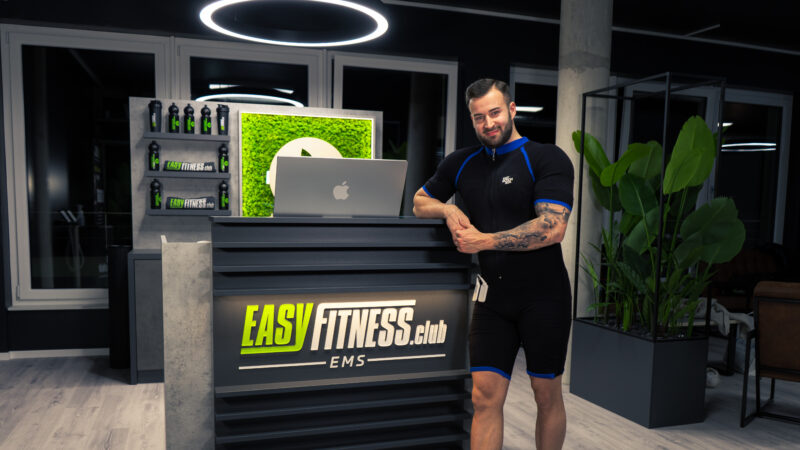 Ein Mann im schwarzen Sportanzug steht lächelnd an der Rezeption eines modernen Fitnessclubs namens EASYFITNESS.club EMS. Im stilvollen, gut beleuchteten Innenraum sind Pflanzen und Fitnessprodukte zu sehen.