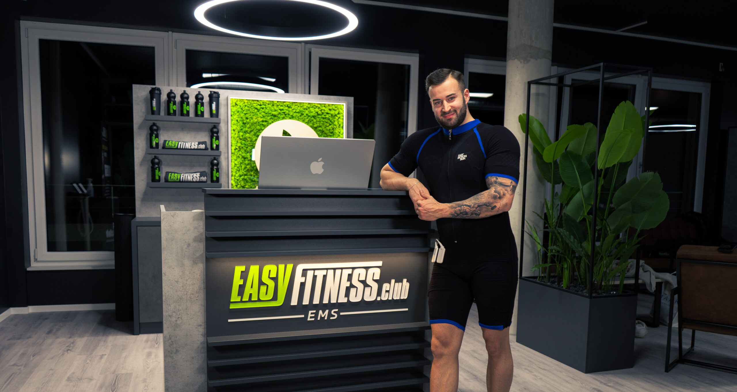 Ein Mann im schwarzen Sportanzug steht lächelnd an der Rezeption eines modernen Fitnessclubs namens EASYFITNESS.club EMS. Im stilvollen, gut beleuchteten Innenraum sind Pflanzen und Fitnessprodukte zu sehen.