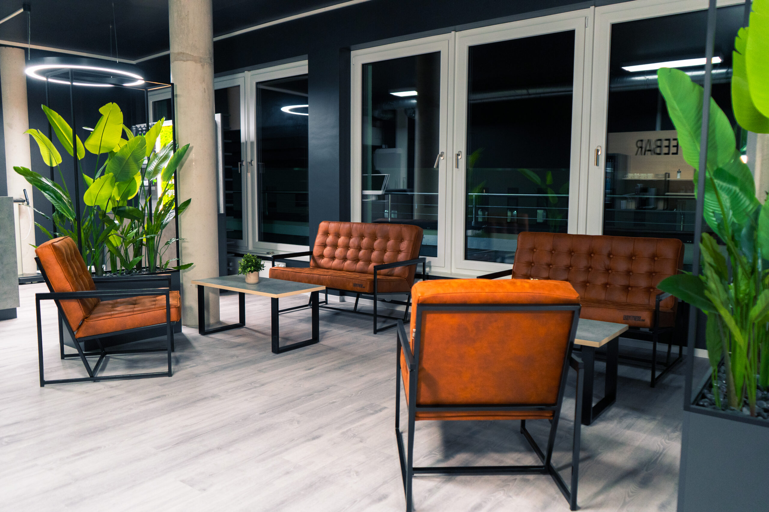 Moderner Loungebereich mit braunen Ledersesseln und Sofas, Holztischen, großen grünen Topfpflanzen und raumhohen Fenstern. Heller, sauberer Raum mit hellem Holzboden und schwarzen Akzenten.