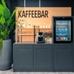 Eine moderne Kaffeebar mit Kaffeemaschine, Gläsern, Tassen und einem kleinen Getränkekühlschrank. Große Grünpflanzen schmücken beide Seiten. Darüber steht ein Holzschild mit der Aufschrift „KAFFEEBAR“. Eine moderne Kaffeebar mit Kaffeemaschine, Gläsern, Tassen und einem kleinen Getränkekühlschrank. Große Grünpflanzen schmücken beide Seiten. Darüber steht ein Holzschild mit der Aufschrift „KAFFEEBAR“.