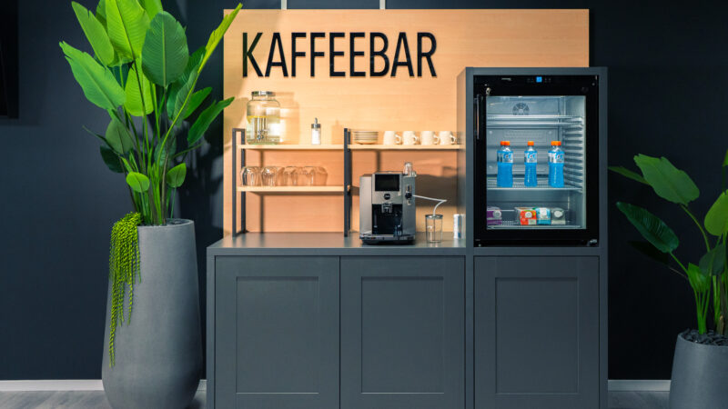 Eine moderne Kaffeebar mit Kaffeemaschine, Gläsern, Tassen und einem kleinen Getränkekühlschrank. Große Grünpflanzen schmücken beide Seiten. Darüber steht ein Holzschild mit der Aufschrift „KAFFEEBAR“.