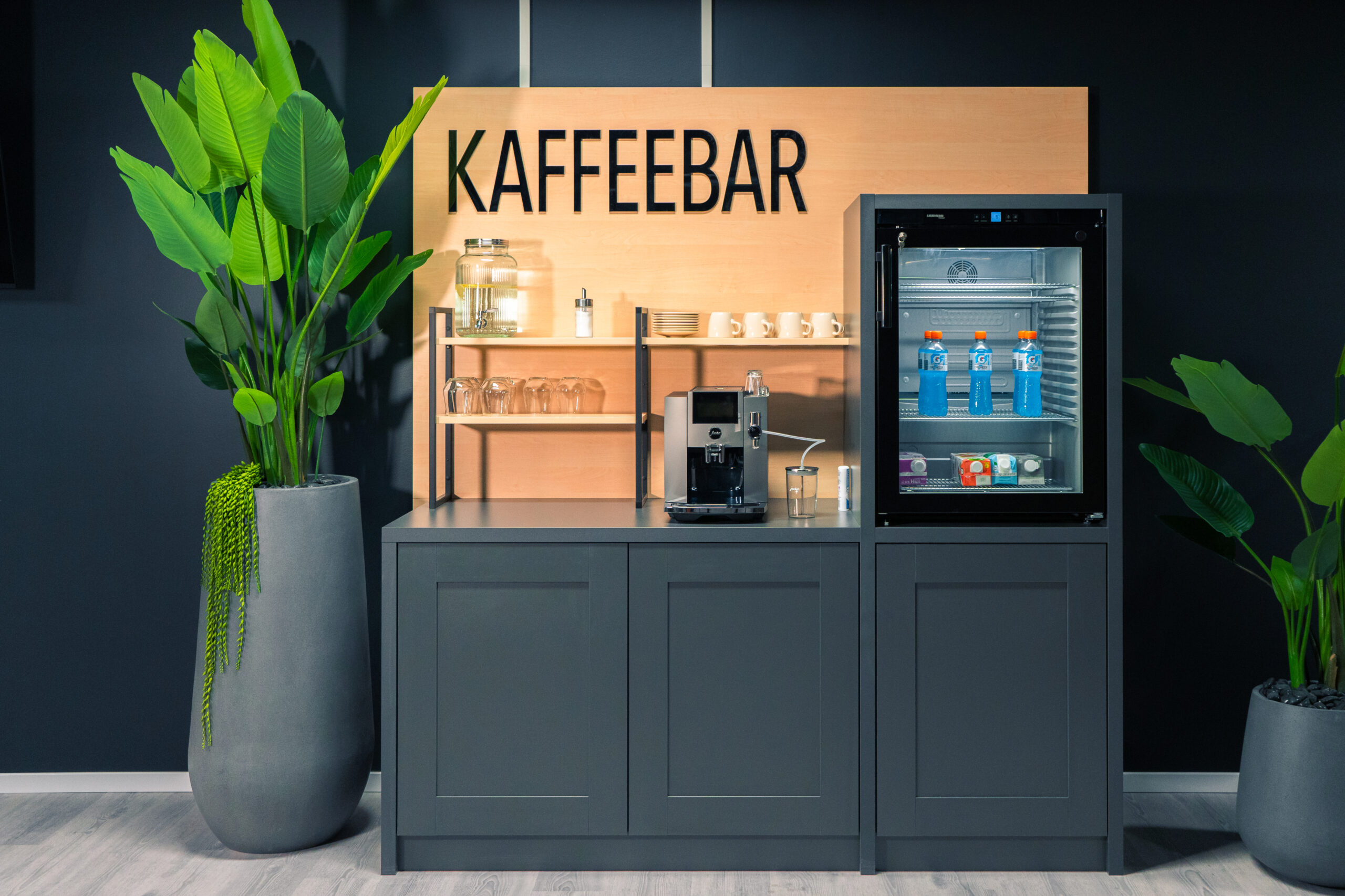 Eine moderne Kaffeebar mit Kaffeemaschine, Gläsern, Tassen und einem kleinen Getränkekühlschrank. Große Grünpflanzen schmücken beide Seiten. Darüber steht ein Holzschild mit der Aufschrift „KAFFEEBAR“.