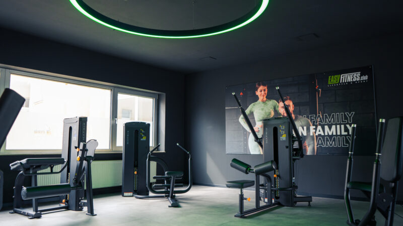 Ein modernes Fitnessstudio mit verschiedenen Trainingsgeräten, großen Fenstern, die viel Tageslicht hereinlassen, und einem Wandposter mit zwei lächelnden Menschen und den Worten „FAMILY“ und „EASY FITNESS“. Der Raum ist sauber und gut beleuchtet.