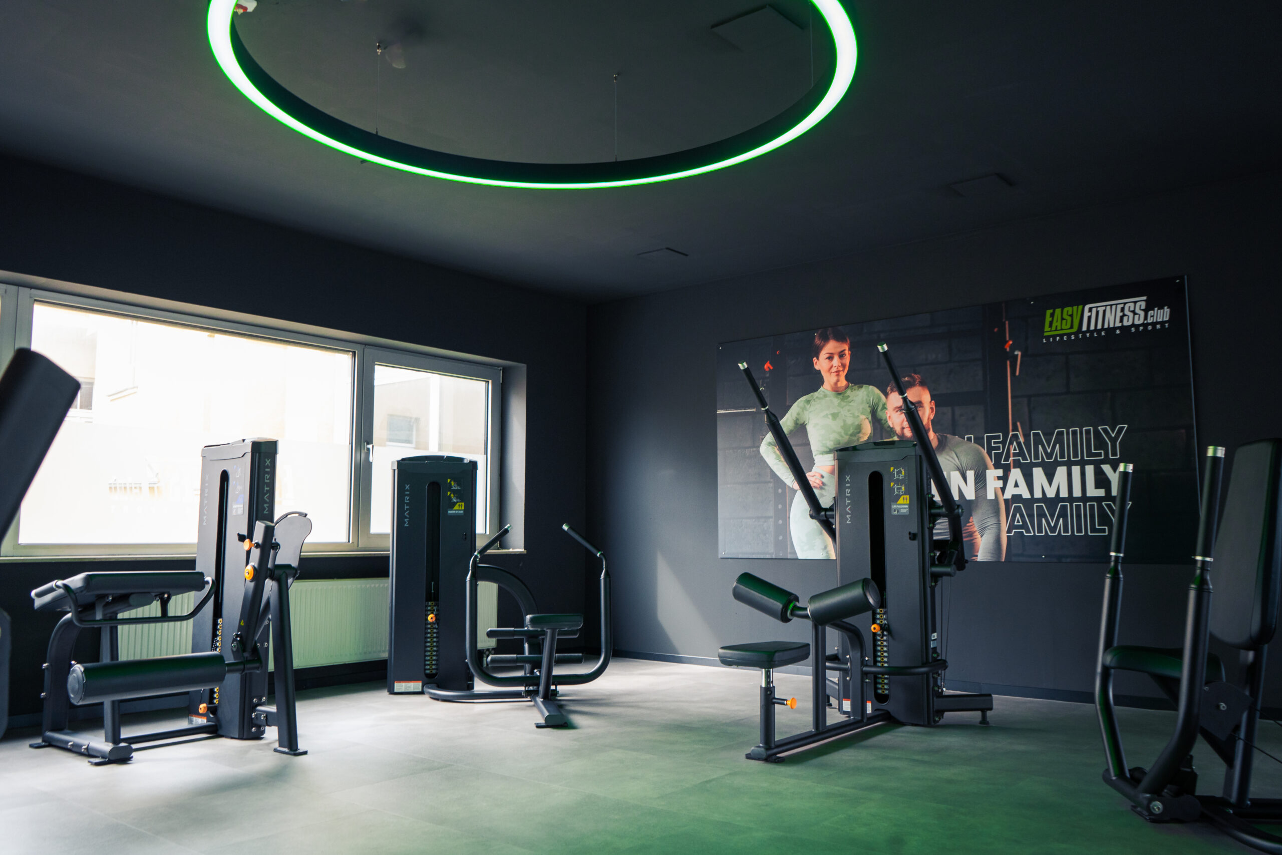 Ein modernes Fitnessstudio mit verschiedenen Trainingsgeräten, großen Fenstern, die viel Tageslicht hereinlassen, und einem Wandposter mit zwei lächelnden Menschen und den Worten „FAMILY“ und „EASY FITNESS“. Der Raum ist sauber und gut beleuchtet.