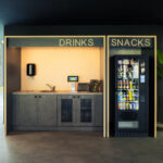 Ein moderner Büro-Snackbereich mit einer Getränkestation auf der linken Seite mit Spüle und Kaffeemaschinen und einem Snackautomaten auf der rechten Seite, beide mit der Aufschrift „Getränke“ und „Snacks“. In der Nähe steht eine grüne Pflanze. Ein moderner Büro-Snackbereich mit einer Getränkestation auf der linken Seite mit Spüle und Kaffeemaschinen und einem Snackautomaten auf der rechten Seite, beide mit der Aufschrift „Getränke“ und „Snacks“. In der Nähe steht eine grüne Pflanze.