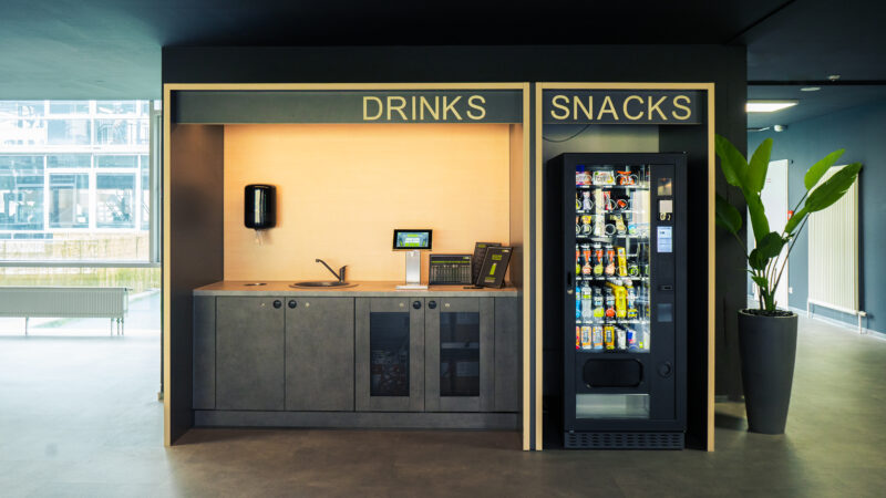 Ein moderner Büro-Snackbereich mit einer Getränkestation auf der linken Seite mit Spüle und Kaffeemaschinen und einem Snackautomaten auf der rechten Seite, beide mit der Aufschrift „Getränke“ und „Snacks“. In der Nähe steht eine grüne Pflanze.