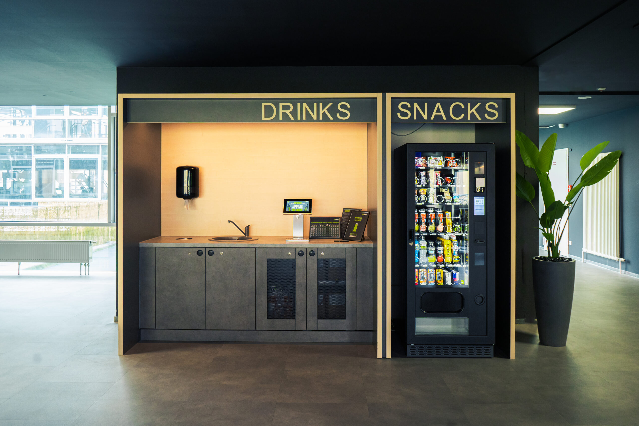 Ein moderner Büro-Snackbereich mit einer Getränkestation auf der linken Seite mit Spüle und Kaffeemaschinen und einem Snackautomaten auf der rechten Seite, beide mit der Aufschrift „Getränke“ und „Snacks“. In der Nähe steht eine grüne Pflanze.