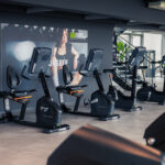 Vier stationäre Heimtrainer stehen in einem modernen Fitnessstudio aufgereiht, dahinter hängt ein großes Motivationsposter. Der Raum ist hell und geräumig, im Hintergrund sind Fitnessgeräte zu sehen. Vier stationäre Heimtrainer stehen in einem modernen Fitnessstudio aufgereiht, dahinter hängt ein großes Motivationsposter. Der Raum ist hell und geräumig, im Hintergrund sind Fitnessgeräte zu sehen.