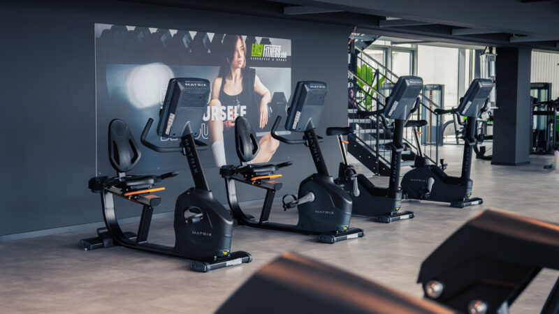 Vier stationäre Heimtrainer stehen in einem modernen Fitnessstudio aufgereiht, dahinter hängt ein großes Motivationsposter. Der Raum ist hell und geräumig, im Hintergrund sind Fitnessgeräte zu sehen.
