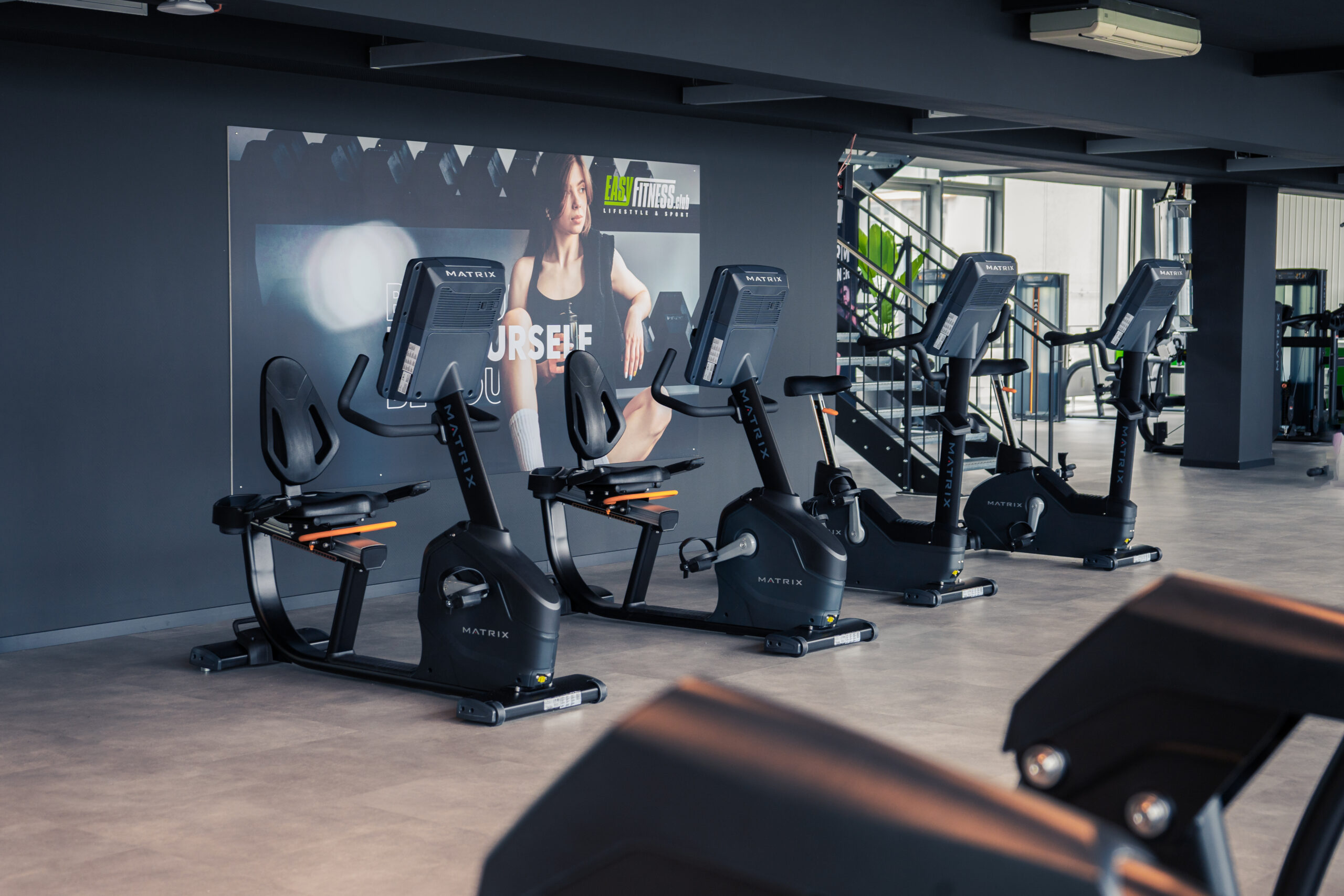 Vier stationäre Heimtrainer stehen in einem modernen Fitnessstudio aufgereiht, dahinter hängt ein großes Motivationsposter. Der Raum ist hell und geräumig, im Hintergrund sind Fitnessgeräte zu sehen.
