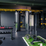 Ein modernes Fitnessstudio mit Kraftgeräten, Kugelhanteln, einem Schlitten und einem funktionellen Trainingsgerät. Der Raum ist mit schwarzen und gelben Akzenten und Gummiböden ausgestattet. Ein modernes Fitnessstudio mit Kraftgeräten, Kugelhanteln, einem Schlitten und einem funktionellen Trainingsgerät. Der Raum ist mit schwarzen und gelben Akzenten und Gummiböden ausgestattet.