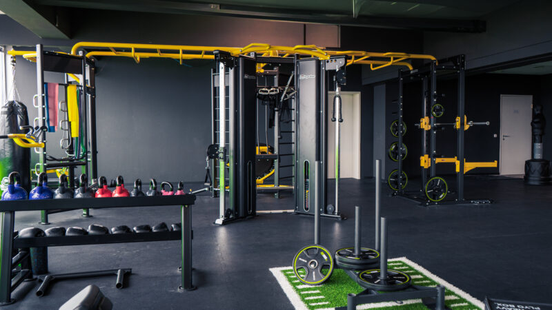 Ein modernes Fitnessstudio mit Kraftgeräten, Kugelhanteln, einem Schlitten und einem funktionellen Trainingsgerät. Der Raum ist mit schwarzen und gelben Akzenten und Gummiböden ausgestattet.