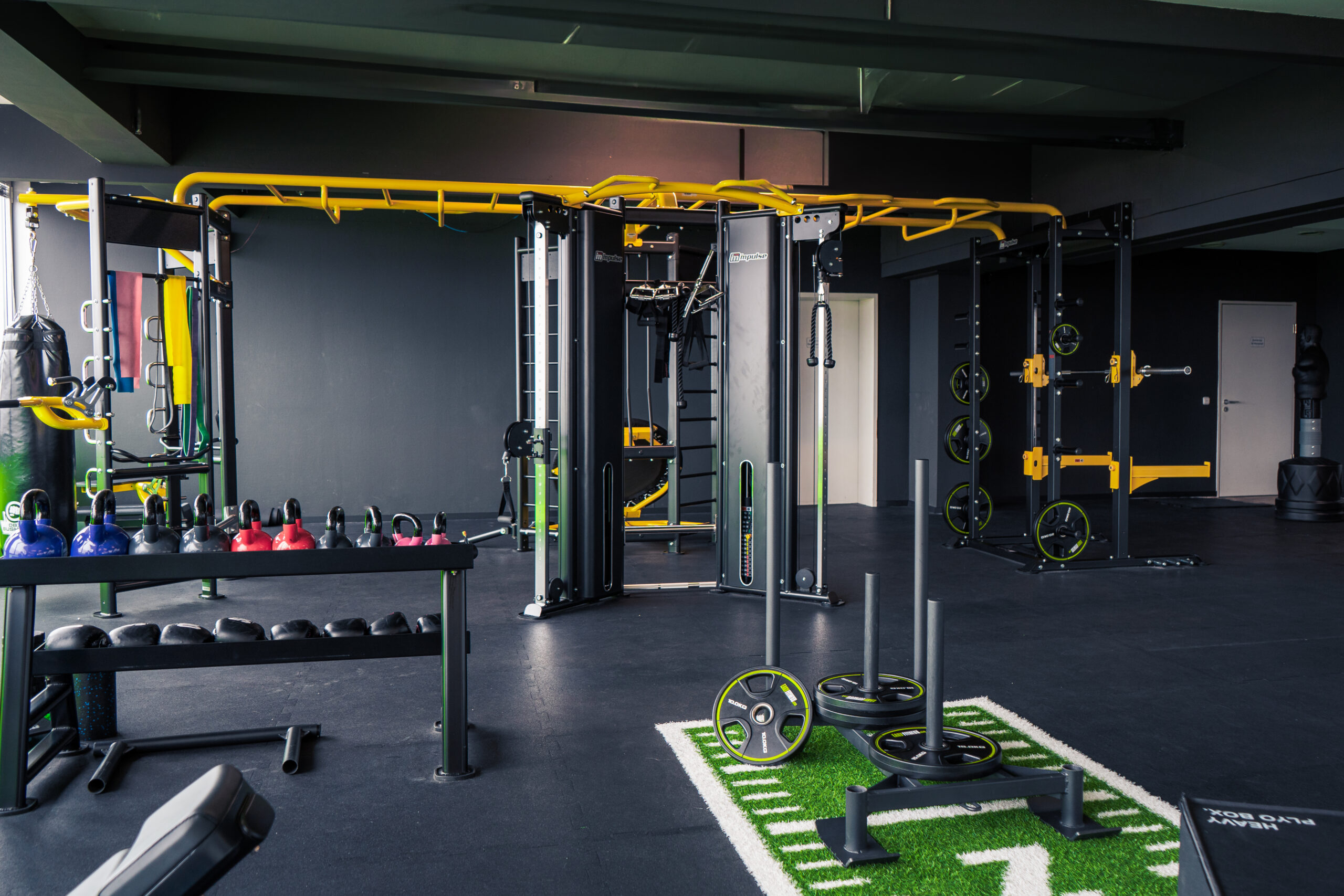 Ein modernes Fitnessstudio mit Kraftgeräten, Kugelhanteln, einem Schlitten und einem funktionellen Trainingsgerät. Der Raum ist mit schwarzen und gelben Akzenten und Gummiböden ausgestattet.