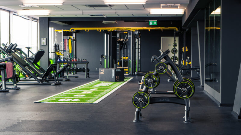 Ein modernes Fitnessstudio mit Gewichthebergeräten, Bänken, Maschinen, einem Hantelständer und einem grünen Kunstrasenstreifen mit weißen Markierungen, alles in einem geräumigen, gut beleuchteten Raum mit schwarzen Böden und Wänden.