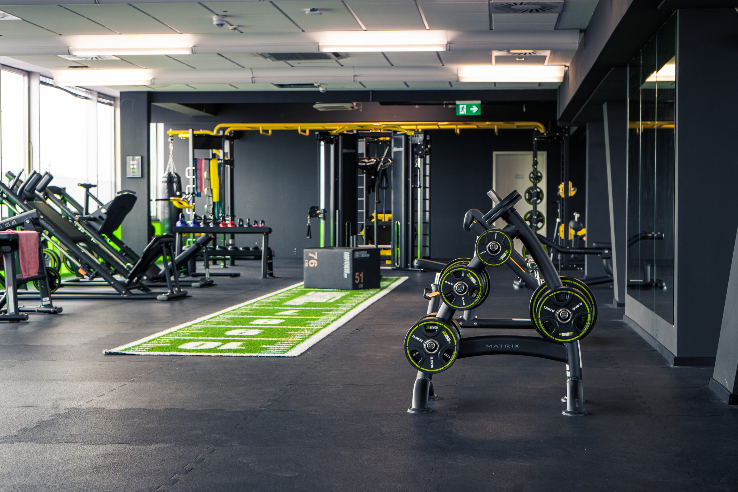 Ein modernes Fitnessstudio mit Gewichthebergeräten, Bänken, Maschinen, einem Hantelständer und einem grünen Kunstrasenstreifen mit weißen Markierungen, alles in einem geräumigen, gut beleuchteten Raum mit schwarzen Böden und Wänden.