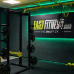 Ein moderner Fitnessraum mit Kraftgeräten, Hanteln und Widerstandsbändern. An der Wand hängt ein Schild mit der Aufschrift „Easy Fitness Club – The Smart Gym“, beleuchtet von grünen LED-Leuchten. Ein moderner Fitnessraum mit Kraftgeräten, Hanteln und Widerstandsbändern. An der Wand hängt ein Schild mit der Aufschrift „Easy Fitness Club – The Smart Gym“, beleuchtet von grünen LED-Leuchten.