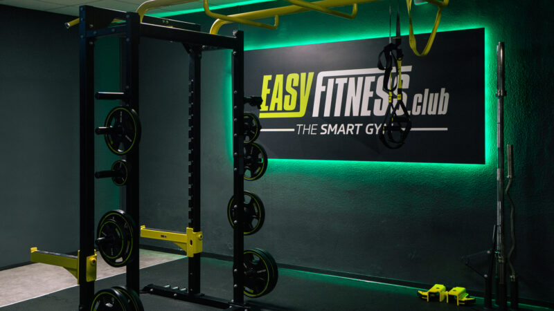 Ein moderner Fitnessraum mit Kraftgeräten, Hanteln und Widerstandsbändern. An der Wand hängt ein Schild mit der Aufschrift „Easy Fitness Club – The Smart Gym“, beleuchtet von grünen LED-Leuchten.