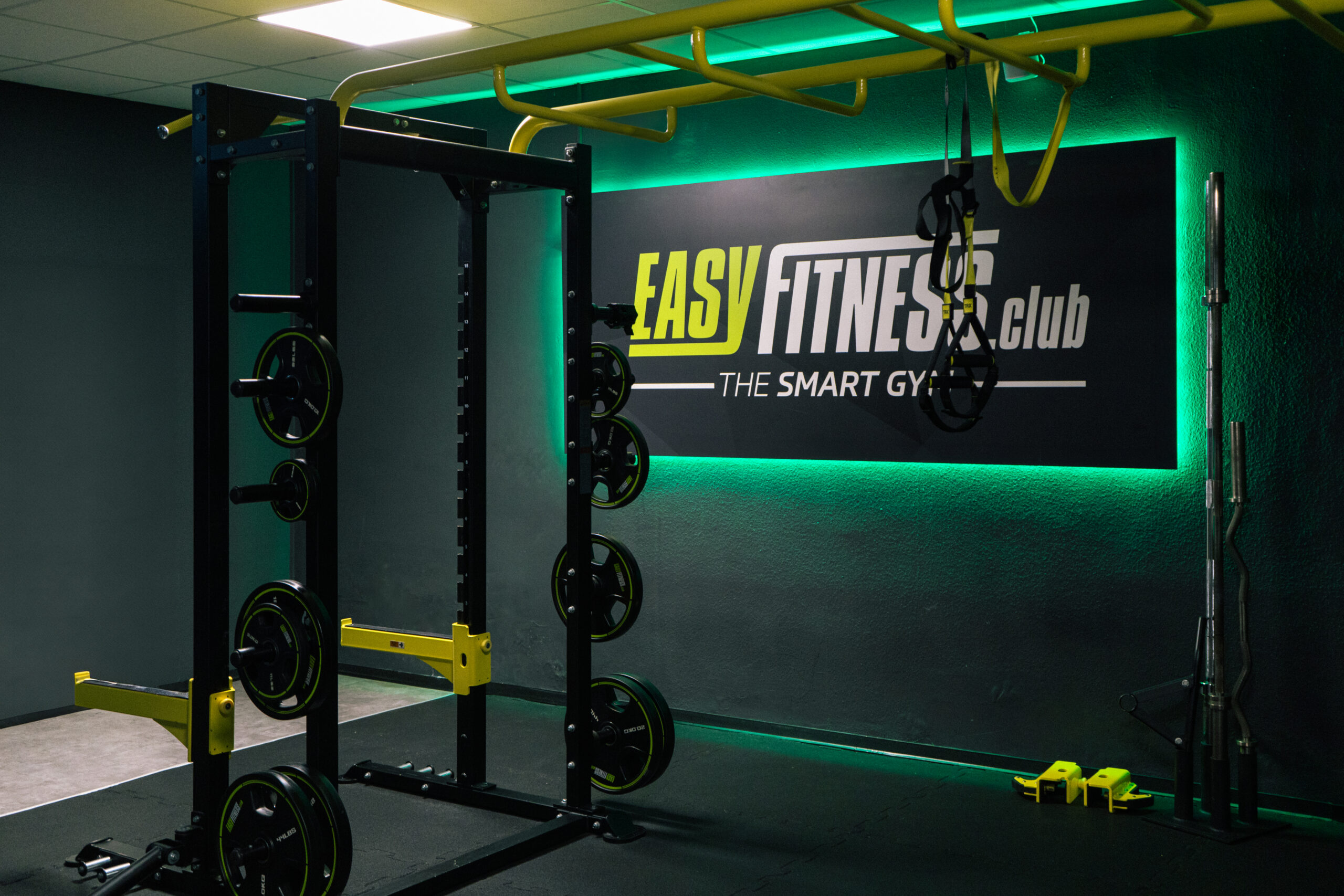 Ein moderner Fitnessraum mit Kraftgeräten, Hanteln und Widerstandsbändern. An der Wand hängt ein Schild mit der Aufschrift „Easy Fitness Club – The Smart Gym“, beleuchtet von grünen LED-Leuchten.