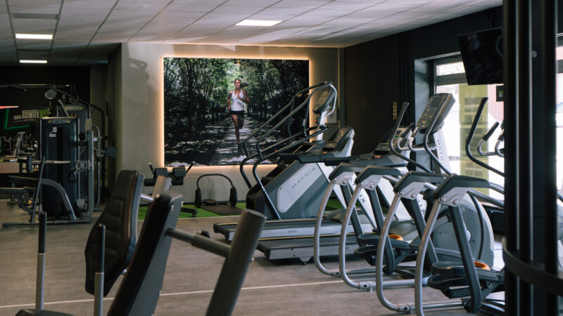 Ein modernes Fitnessstudio mit mehreren Laufbändern und Trainingsgeräten. An der Wand hängt ein großes Foto einer Person, die im Wald joggt. Das Fitnessstudio ist gut beleuchtet – sowohl mit natürlichem als auch mit künstlichem Licht.