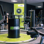 Ein modernes Fitnessstudio mit verschiedenen Trainingsgeräten und einem schwarzen Fass mit dem „EASYFITNESS“-Logo in der Mitte. Das Fitnessstudio hat dunkle Wände, eine kreisförmige Bodengrafik in Grün und ein Werbebanner im Hintergrund. Ein modernes Fitnessstudio mit verschiedenen Trainingsgeräten und einem schwarzen Fass mit dem „EASYFITNESS“-Logo in der Mitte. Das Fitnessstudio hat dunkle Wände, eine kreisförmige Bodengrafik in Grün und ein Werbebanner im Hintergrund.