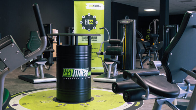 Ein modernes Fitnessstudio mit verschiedenen Trainingsgeräten und einem schwarzen Fass mit dem „EASYFITNESS“-Logo in der Mitte. Das Fitnessstudio hat dunkle Wände, eine kreisförmige Bodengrafik in Grün und ein Werbebanner im Hintergrund.