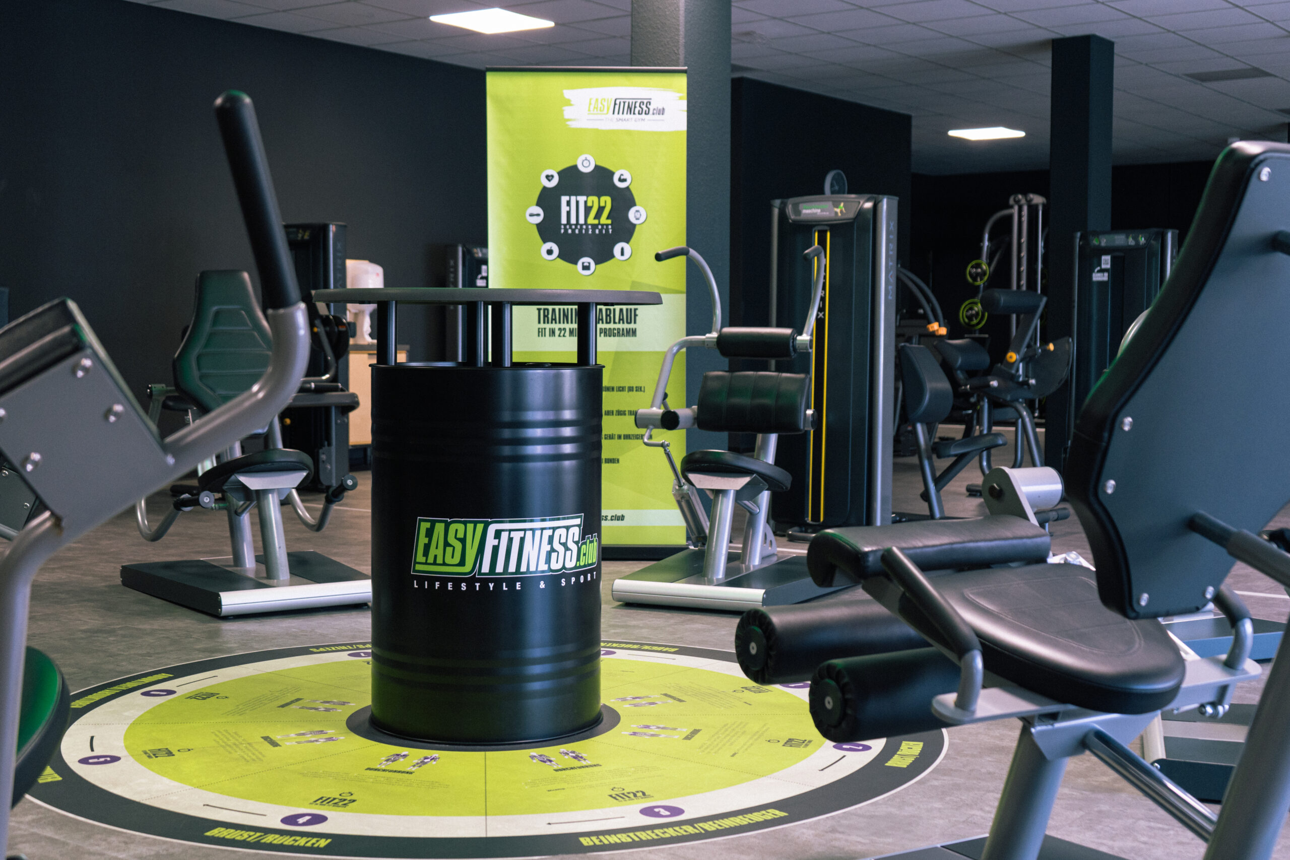 Ein modernes Fitnessstudio mit verschiedenen Trainingsgeräten und einem schwarzen Fass mit dem „EASYFITNESS“-Logo in der Mitte. Das Fitnessstudio hat dunkle Wände, eine kreisförmige Bodengrafik in Grün und ein Werbebanner im Hintergrund.