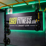 Eine Turnhallenwand mit einem Schild mit der Aufschrift „EASY FITNESS.club THE SMART GYM“, Hantelscheiben auf einem Ständer, gelben Stangen und hängenden Trainingsgurten. Grüne LED-Leuchten beleuchten den Raum. Eine Turnhallenwand mit einem Schild mit der Aufschrift „EASY FITNESS.club THE SMART GYM“, Hantelscheiben auf einem Ständer, gelben Stangen und hängenden Trainingsgurten. Grüne LED-Leuchten beleuchten den Raum.