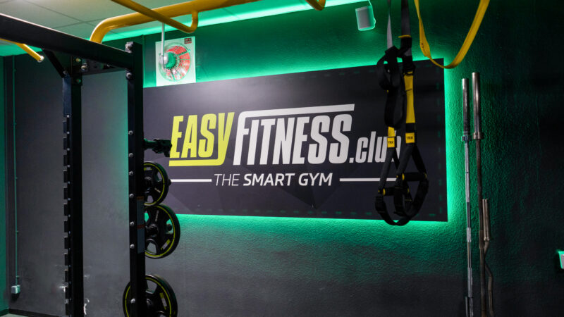Eine Turnhallenwand mit einem Schild mit der Aufschrift „EASY FITNESS.club THE SMART GYM“, Hantelscheiben auf einem Ständer, gelben Stangen und hängenden Trainingsgurten. Grüne LED-Leuchten beleuchten den Raum.