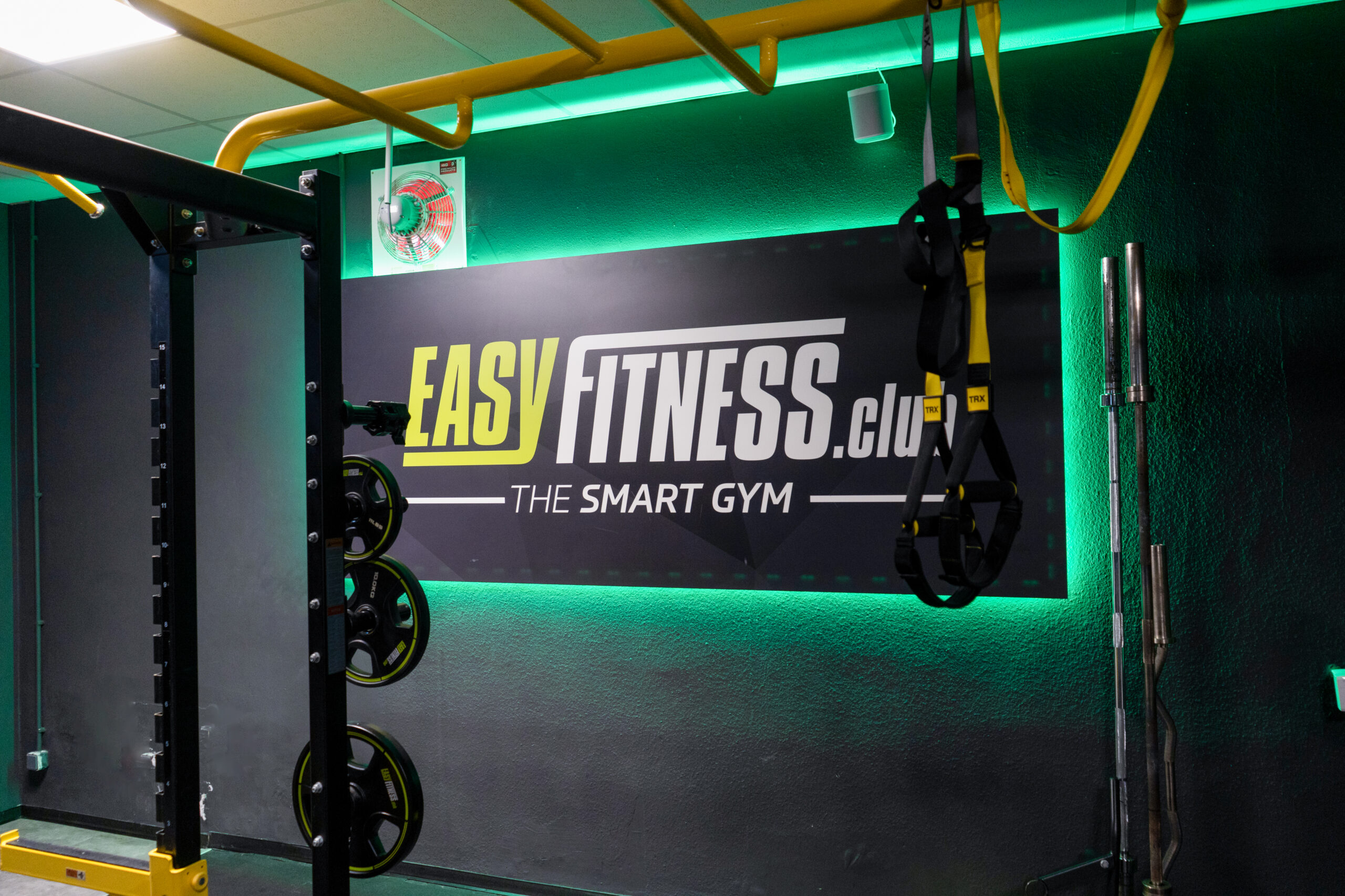 Eine Turnhallenwand mit einem Schild mit der Aufschrift „EASY FITNESS.club THE SMART GYM“, Hantelscheiben auf einem Ständer, gelben Stangen und hängenden Trainingsgurten. Grüne LED-Leuchten beleuchten den Raum.